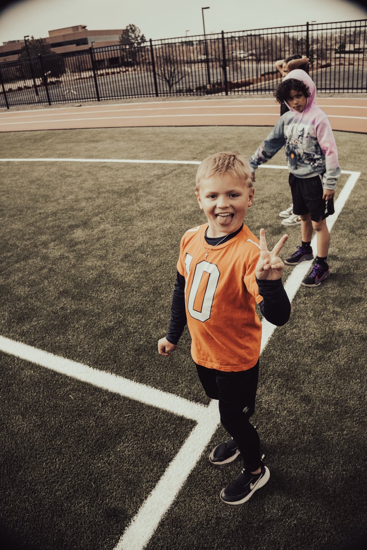 A smiling young athlete in an orange jersey flashing a peace sign on a turf field
